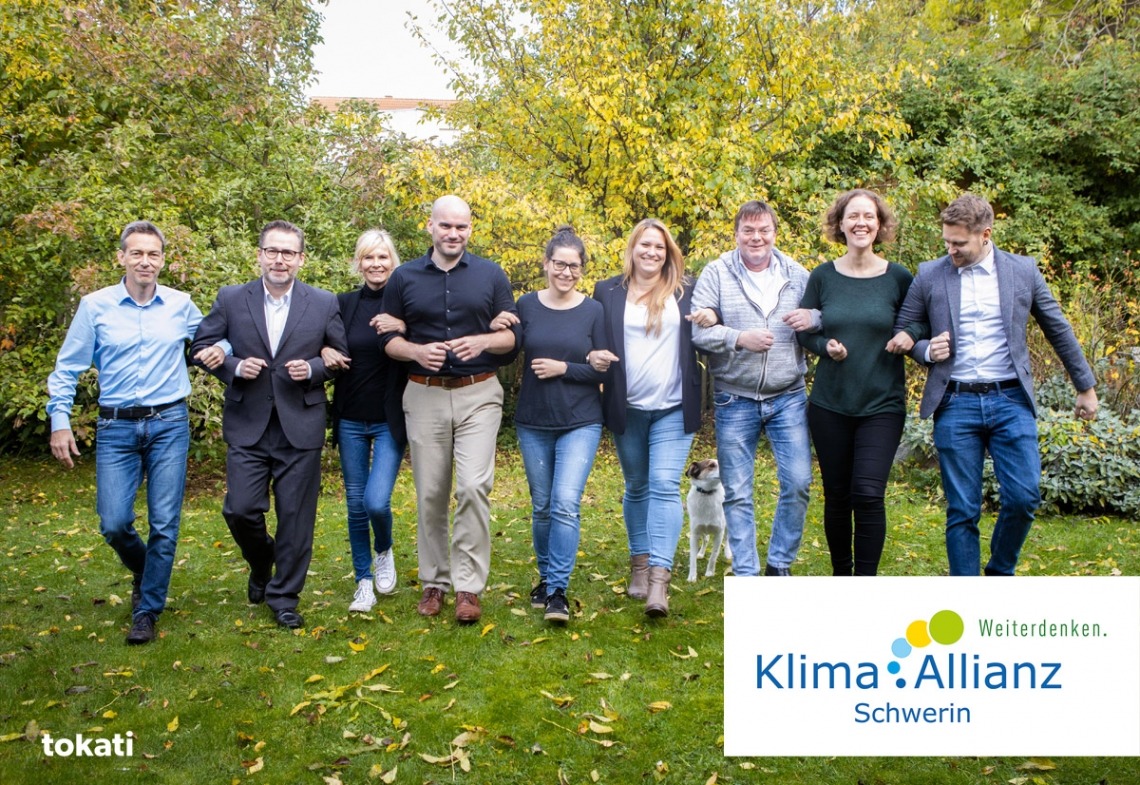Acht Personen gehen Arm in Arm über eine Wiese vor Büschen, symbolisieren Zusammenarbeit für Klima-Allianz Schwerin